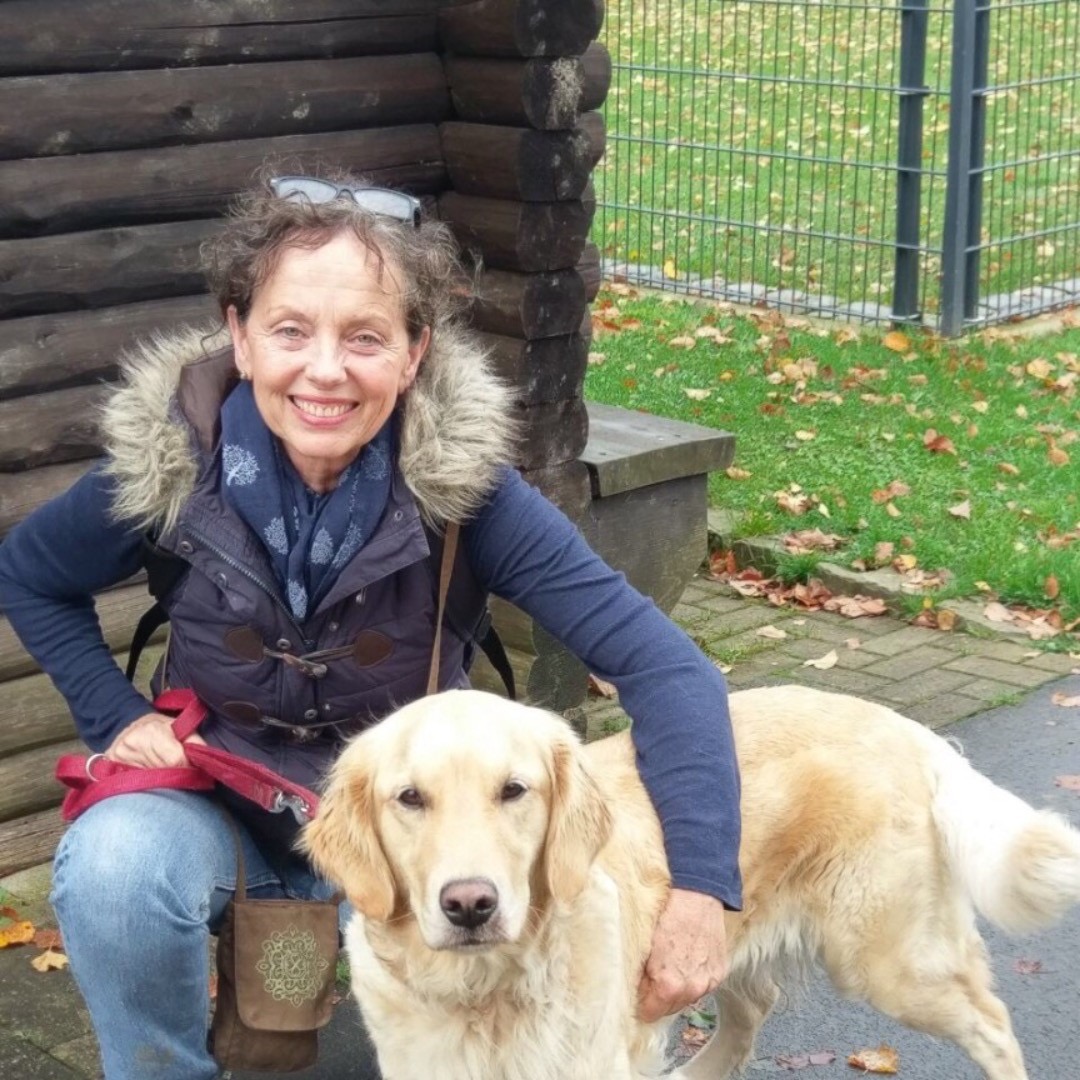 Gabriele kniet draußen mit ihrem Golden Retriever und lächelt in die Kamera.